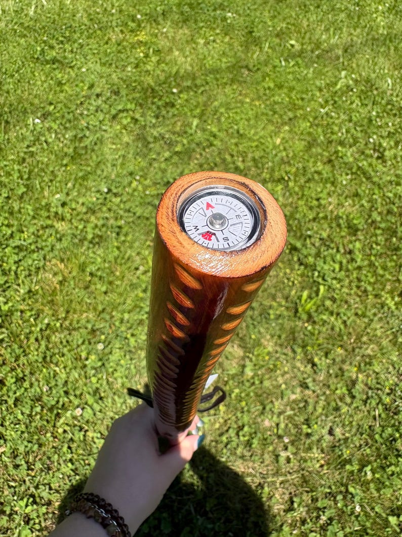 May include: A wooden walking stick with a compass embedded in the top. The stick is brown with a carved design and a black strap. The compass has a white face with black and red markings.