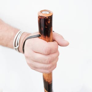 Pu&ograve; includere: Una mano che tiene un bastone da passeggio in legno con finitura marrone e nera. Il bastone ha un cinturino in pelle e un tappo in metallo in cima.