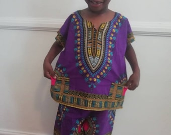 Puede incluir: Un niño con un conjunto dashiki morado. La camiseta de manga corta y los pantalones cortos presentan un diseño estampado colorido con detalles dorados, azules y turquesas. El atuendo es de estilo africano tradicional.