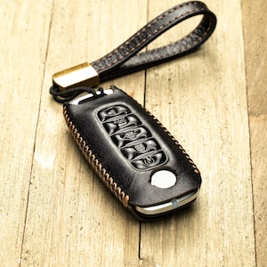 May include: Black leather car key fob cover with a brown stitched edge and a matching wrist strap. The fob has several buttons and a white circular button. The wrist strap has a gold-colored metal clasp.