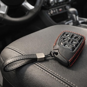 May include: Black leather car key fob cover with red stitching. The key fob has multiple buttons and a black leather wrist strap with a silver metal accent. The key fob is resting on a black leather car seat.