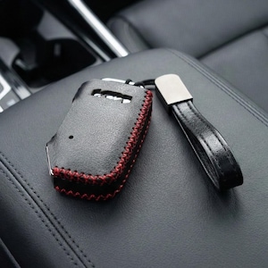 May include: Black leather key fob cover with red stitching and a black leather strap. The key fob cover has a metal accent and is designed to protect car keys. The key fob is resting on a black leather surface.
