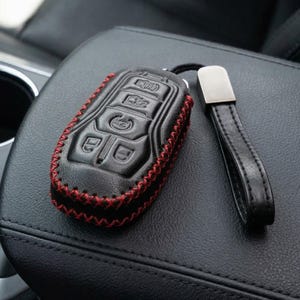 May include: Black leather key fob cover with red stitching. The cover has embossed buttons and a matching black leather strap with a silver-tone metal accent. The key fob cover is designed to protect the car key.