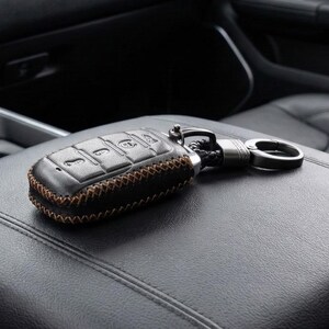 May include: Black leather car key fob cover with brown stitching. The key fob has a metal clasp and a braided leather strap. The key fob is resting on a black leather surface.