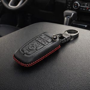 May include: Black leather car key fob cover with red stitching. The key fob has embossed buttons and a metal keyring attachment. The cover is designed to protect the key fob and add a stylish touch.