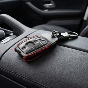 May include: Black leather car key fob cover with red stitching. The key fob has several buttons and a metal keyring attachment. The cover is resting on a black leather surface inside a car.