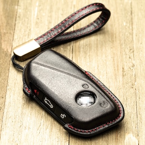 May include: Black leather key fob with a red stitching detail and a gold metal loop. The fob has a car icon and a button.