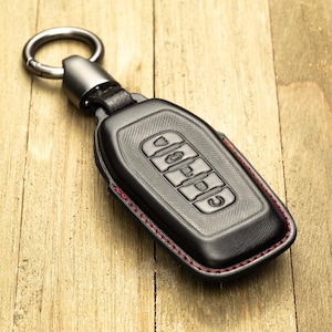 May include: Black leather key fob cover with red stitching and a silver key ring. The cover has embossed buttons and the word "JEEP". The key fob is on a wooden surface.