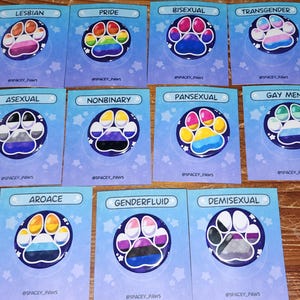 May include: A collection of colorful paw print designs on cards, each representing a different LGBTQ+ identity. The cards feature text such as "Lesbian," "Pride," "Bisexual," and "Transgender." The designs are set against a light blue background with star accents.