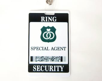 May include: A white plastic badge with black text and a green logo. The badge reads "RING SPECIAL AGENT SECURITY" and features a green shield with two interlocking rings and a heart.