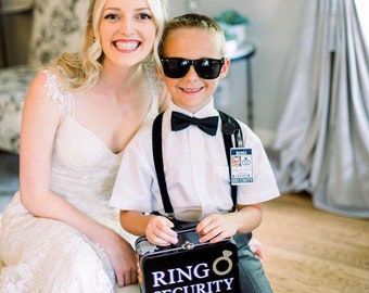 May include: A young boy wearing a white shirt, black bow tie, and sunglasses holds a black metal box with the text "RING SECURITY" on it. He is standing next to a woman wearing a white wedding dress.