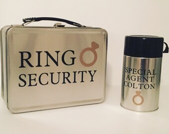May include: A silver lunchbox with the text "RING SECURITY" and a gold ring symbol. Next to it is a silver thermos with the text "SPECIAL AGENT COLTON" and a gold ring symbol.