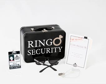 May include: A black metal lunchbox with the words "RINGO SECURITY" printed on the front. The lunchbox is open and there is a black pair of sunglasses inside. There is also a white card with the words "TOP SECRET YOUR MISSION" printed on it. The card is next to a white lanyard with a black ID card that says "SPECIAL AGENT RILEY SECURITY" and has a picture of a person on it.