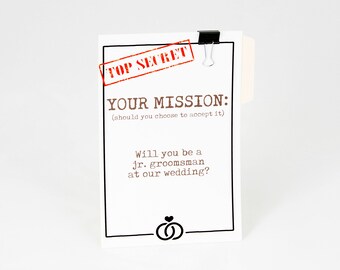 May include: A white card with black text and a red "TOP SECRET" stamp. The card reads "YOUR MISSION: (should you choose to accept it) Will you be a jr. groomsman at our wedding?" with a black and white illustration of two wedding rings.
