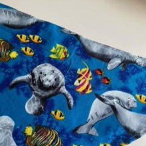 May include: A blue fabric with a pattern of gray manatees and colorful fish. The fish are yellow, orange, and red. The manatees are swimming in the blue water.