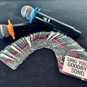 A colorful deck of cards for a game are fanned out. The prominent one reads "Sing your goodbye song". Two karaoke microphones are in the background.