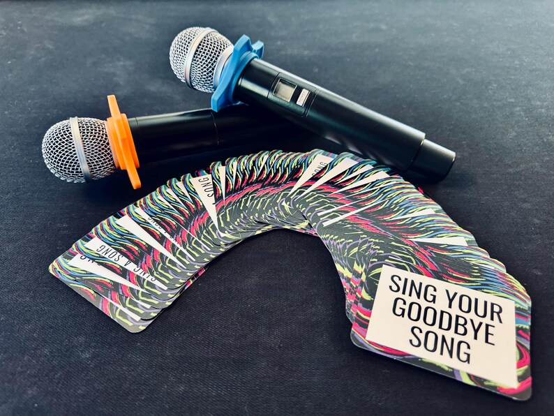 A colorful deck of cards for a game are fanned out. The prominent one reads "Sing your goodbye song". Two karaoke microphones are in the background.