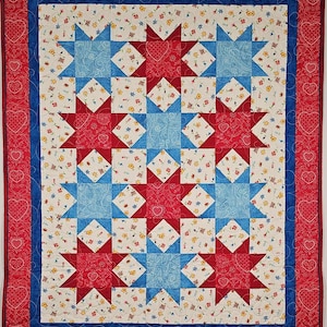 May include: A handmade quilt with a star pattern in red, white, and blue. The quilt features a central design of alternating red and blue stars, surrounded by a white floral pattern. The border is red with heart shapes.