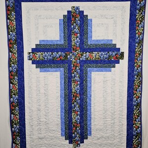 May include: A rectangular quilt featuring a central cross design constructed from fabric strips in shades of blue and a floral print with blue and red flowers. The quilt has a white background with quilted patterns and a blue floral border.