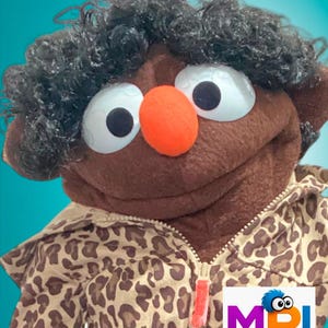 May include: A brown puppet with curly black hair, large white eyes with black pupils, and a bright orange nose. The puppet wears a tan and brown leopard print zip-up jacket. A logo in the corner reads MPJ MY PUPPET JOURNEY with a blue-haired character.