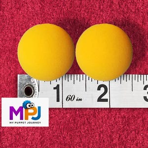 May include: Two yellow foam balls, each approximately 2 inches in diameter, are displayed on a red textured surface. A measuring tape is placed below the balls, and a logo for "My Puppet Journey" is in the lower left corner.