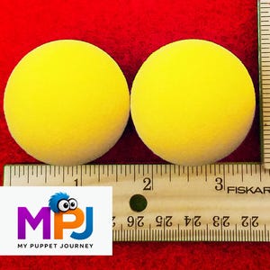 May include: Two bright yellow foam balls are displayed on a red surface next to a ruler. The balls are round and appear to be about 3 inches in diameter. The logo "MPJ MY PUPPET JOURNEY" is visible in the lower left corner.