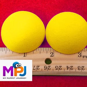 May include: Two bright yellow foam balls, each approximately 2 inches in diameter, are displayed on a red surface next to a ruler. The balls have a textured surface. The logo for "My Puppet Journey" is in the lower left corner.