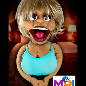 May include: A handmade puppet with a blonde wig, large open mouth, and a blue top. The puppet has a light brown complexion and is set against a blurred background. The puppet's mouth is open, revealing a pink tongue.