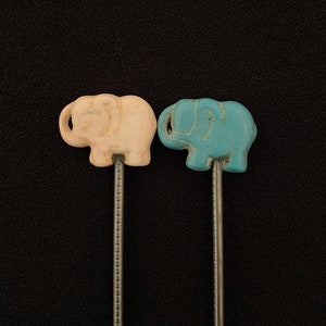 May include: Two small elephant figurines on metal sticks. The elephant on the left is light pink and the elephant on the right is light blue.