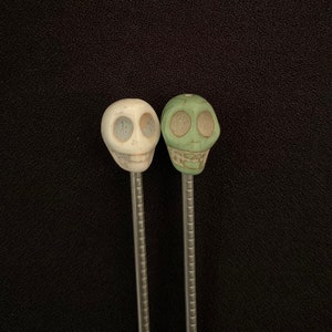 May include: Two skull-shaped beads on silver metal rods. The skull on the left is white, and the skull on the right is green.