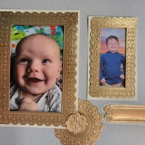 May include: A collection of gold-colored picture frames with scalloped edges, holding photos of a baby and a young child. Additional decorative elements include a round embellishment and a small rectangular frame, all in matching gold tones.