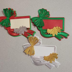 May include: Three Christmas-themed gift tag ornaments. Each features a ribbon and bow design in red, green, silver, and gold. The tags have space for writing and are decorated with holly leaves.