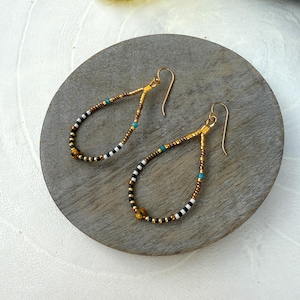 May include: A pair of teardrop-shaped earrings with gold-colored ear wires. The earrings are beaded with a pattern of gold, black, white, and turquoise beads. The earrings are displayed on a gray, circular stone.