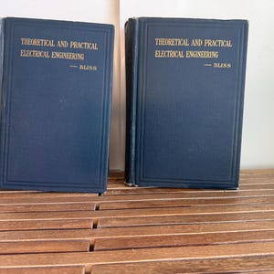 May include: Two navy blue books with gold lettering. The text on the cover reads "THEORETICAL AND PRACTICAL ELECTRICAL ENGINEERING - BLISS". The books are standing upright on a wooden surface.