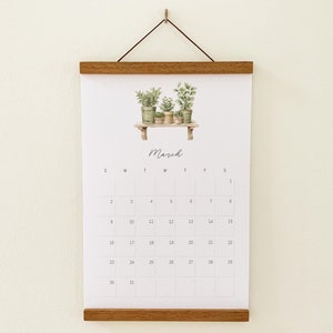 Puede incluir: Un calendario blanco con un marco de madera colgado en una pared blanca. El calendario presenta una ilustración de acuarela de plantas en macetas en un estante y el mes de marzo. La cuadrícula del calendario muestra los días del mes.