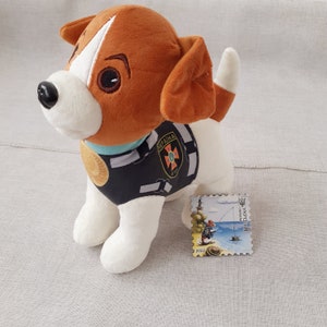 May include: A plush toy dog wearing a black and white uniform with the word "UKRAINE" and a blue and white cross on the chest. The dog is sitting on a white surface with a postage stamp featuring a cartoon dog fishing.