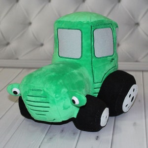 以下が含まれることがあります： 緑のぬいぐるみトラクター。黒い車輪と白いアクセントが付いています。トラクターは大きな目で笑顔をしています。