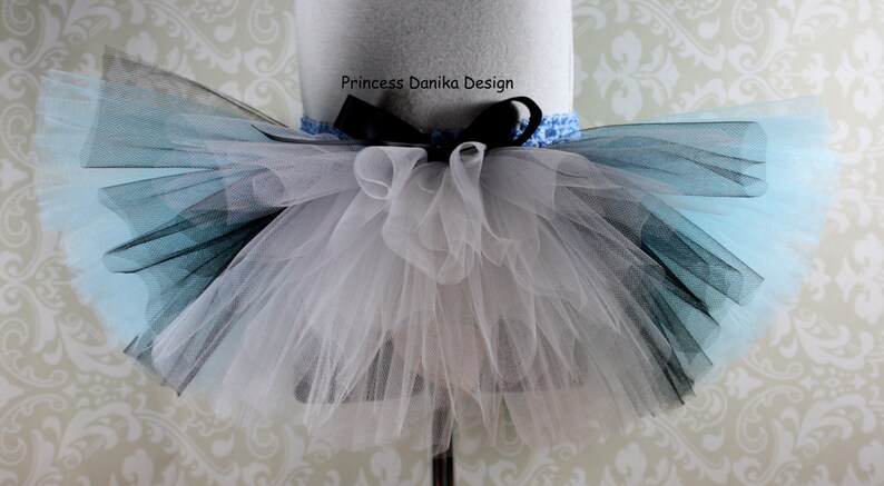 May include: A white, black, and light blue tulle tutu with a black ribbon and bow. The tutu is on a white mannequin. The text "Princess Danika Design" is on the ribbon.