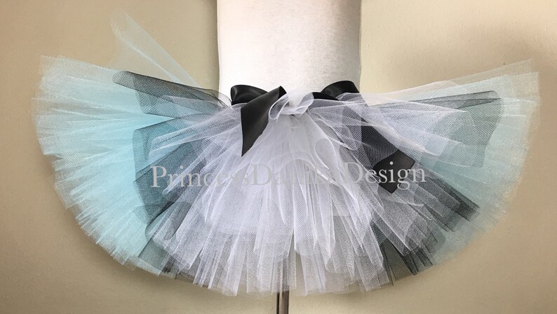 May include: A multi-layered tulle tutu skirt with a black satin bow. The skirt is ombre, starting with light blue, then black, and finally white.