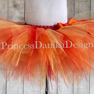 May include: A multi-layered tulle tutu skirt in shades of orange, yellow, and red. The skirt has a satin ribbon waistband and is perfect for a fall-themed costume or party.