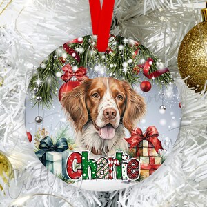 May include: Round Christmas ornament featuring a watercolor portrait of a dog with brown and white fur. The ornament has a red ribbon for hanging and is decorated with Christmas elements, including gifts and greenery. The dog's name, "Charlie", is displayed in festive lettering.