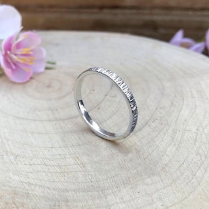 May include: A simple silver band ring with a textured finish. The ring is shown on a wooden surface.