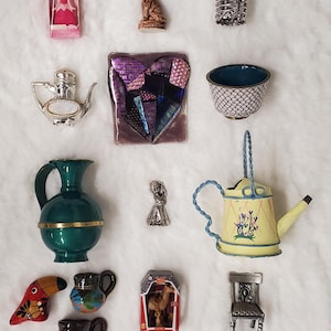 May include: A collection of miniature decorative items, including a doll in a pink box, a cat figurine, a birdcage, a silver teapot, a heart-shaped mosaic, a small bowl, a green pitcher, a silver figurine, a yellow watering can, a toucan, and miniature mugs.