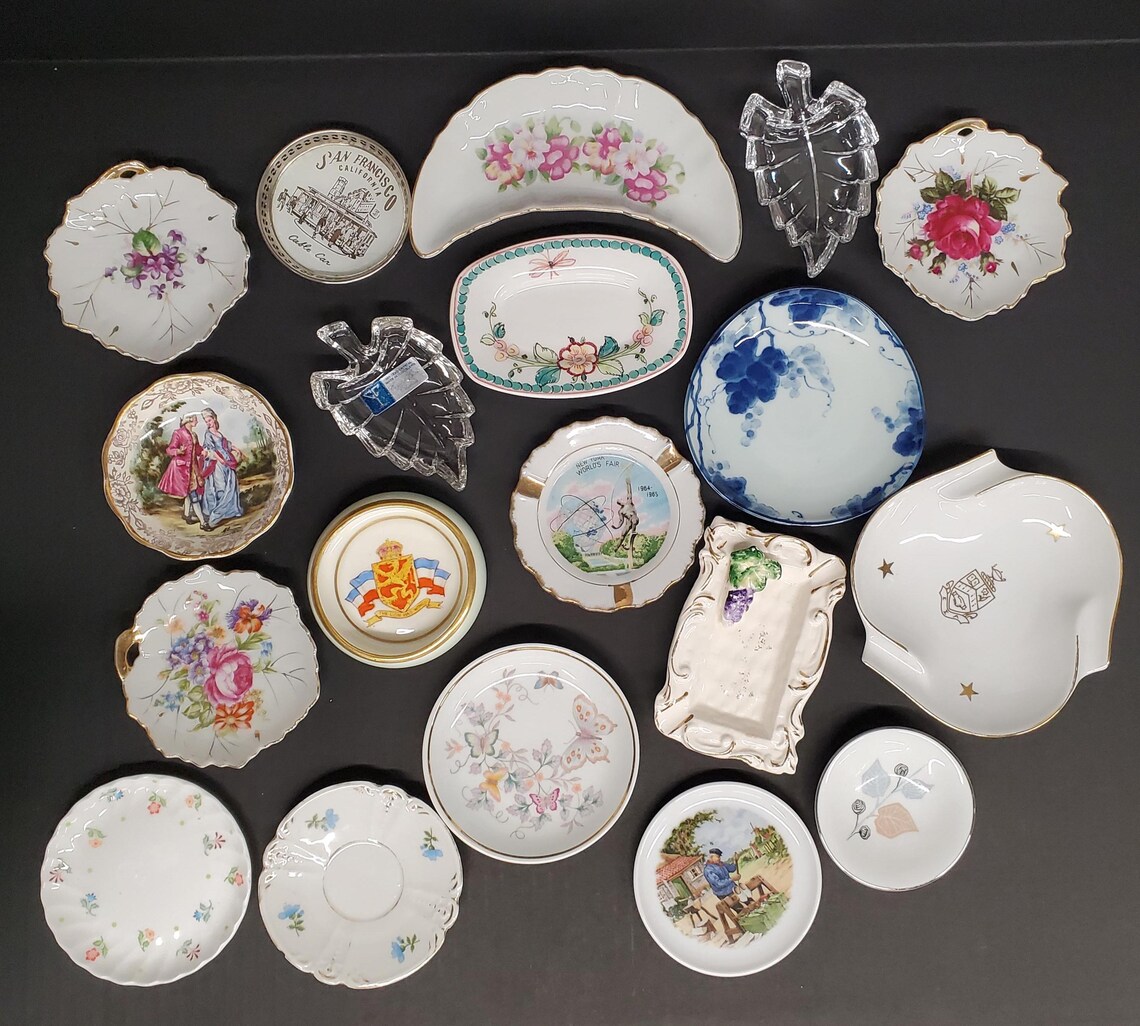 May include: A collection of decorative plates and dishes in various shapes and sizes. The plates feature floral, butterfly, and scenic designs, with gold trim and a blue and white grape pattern. Some plates have handles or leaf-shaped designs.