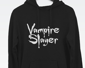 Könnte beinhalten: Ein schwarzer Kapuzenpullover mit dem weißen Schriftzug Vampire Slayer auf der Brust. Der Hoodie verfügt über eine Kängurutasche auf der Vorderseite, lange Ärmel und eine Kapuze mit Kordelzug. Das Kleidungsstück hängt an einem schwarzen Bügel.