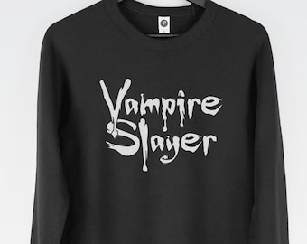Könnte beinhalten: Ein schwarzes Langarm-Sweatshirt mit dem weißen Schriftzug Vampire Slayer auf der Vorderseite. Das Kleidungsstück hängt an einem schwarzen Kleiderbügel und zeigt ein WRAP-Zertifizierungslogo, ein PETA-Approved Vegan-Logo und das Geekdown Apparel-Logo.