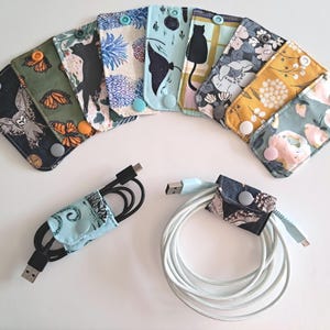 May include: A collection of fabric cable organizers in various patterns, including owls, butterflies, and floral designs. Each organizer has a snap closure. Two organizers hold black and white cables.