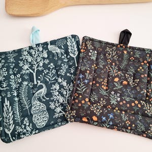 Puede incluir: Dos agarraderas cuadradas acolchadas. Una es de color verde azulado con un estampado de pavo real y flores blancas. La otra es negra con un estampado floral en naranja, azul y blanco. Cada una tiene un lazo de tela para colgar.
