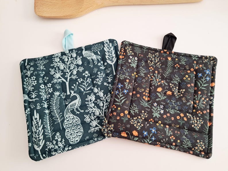 Puede incluir: Dos agarraderas cuadradas acolchadas. Una es de color verde azulado con un estampado de pavo real y flores blancas. La otra es negra con un estampado floral en naranja, azul y blanco. Cada una tiene un lazo de tela para colgar.