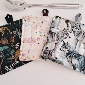 May include: Three square pot holders with hanging loops. One features a black background with a leopard and tropical foliage. Another has a floral pattern with a sleeping deer. The third displays a wolf pack.